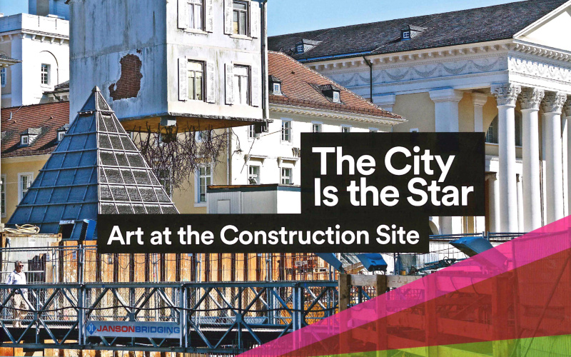 Blick auf den Marktplatz der Stadt Karlsruhe: An einem Kran hängt ein Haus, aus dessen Boden Wurzeln sprießen