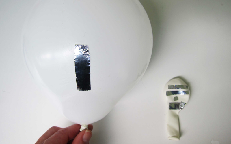 Ein aufgeblasener weißer Ballon mit einem Streifen glänzenden metallenen Klebeband auf weißem Grund. Daneben ein noch nicht aufgeblasener Ballon mir vielen solcher Klebestreifen.