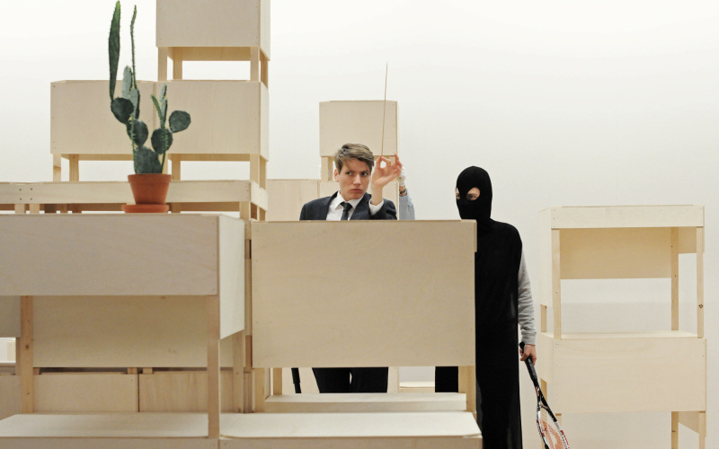 With a construction of light wooden furniture-sculptures are two persons of undefined gender. They are looking at a cactus.