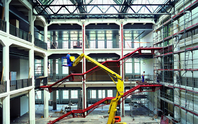 Das Foto zeigt eine historische Ansicht des Umbaus des alten MNK Hallenbaus. Ein imposanter großer gelber Bagger steht mitten in der Halle während im Hintergrund die Rohkonstruktion der zukünftigen Treppen des Ausstellungsraumes in rot hell leuchten.