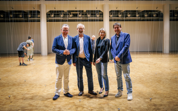 3 Männer, darunter der Kulturbürgermeister und der Vorstand des ZKM, sowie eine Frau, die Kulturamtsleiterin der Stadt Karlsruhe stehen im Lichthof. Dahinter sind die Pendel von William Forsythe zu sehen. 